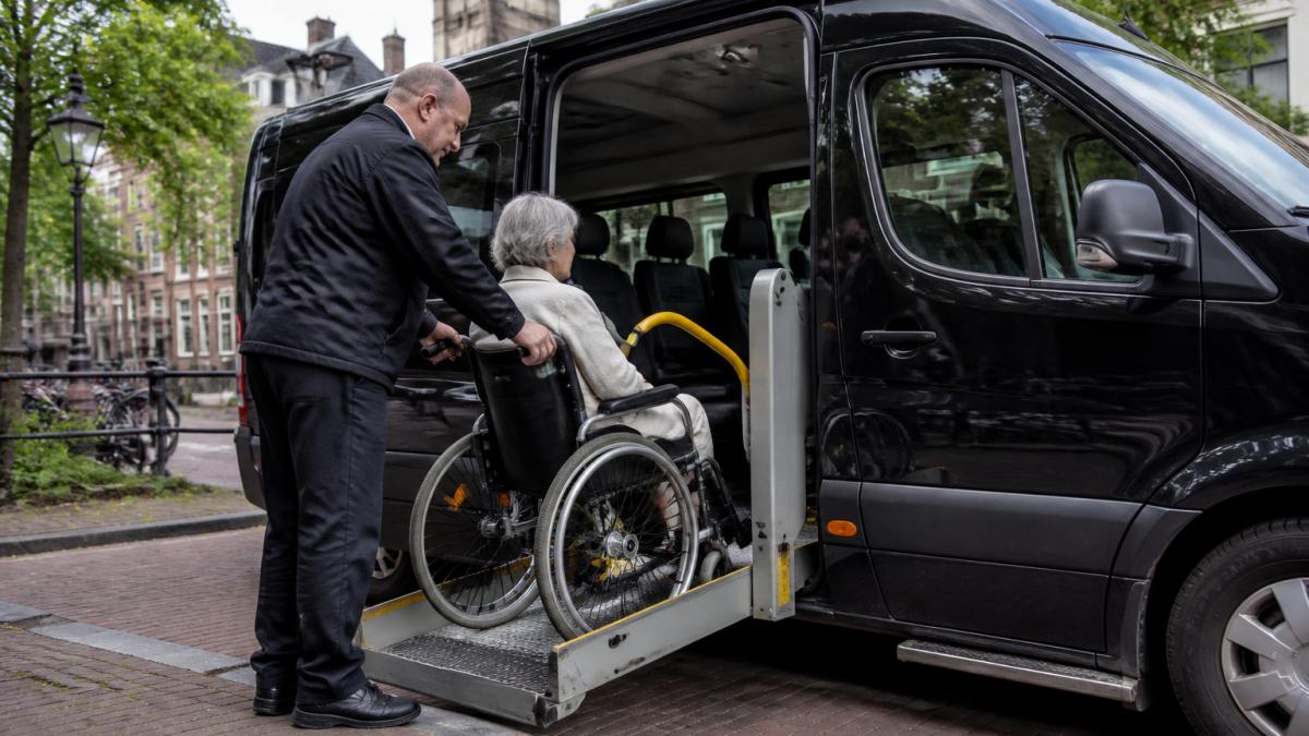 Chauffeur helpt oudere vrouw in rolstoeltaxi voor medische afspraak boeken via de lift in Utrecht.