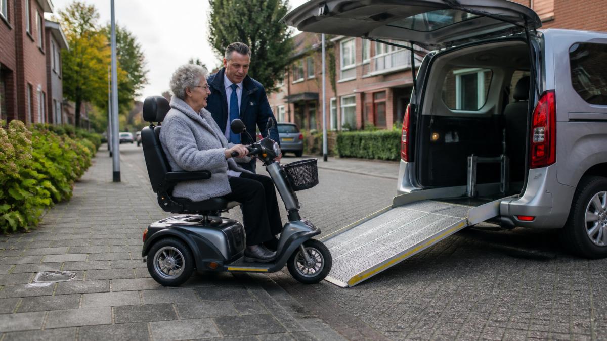 Een vriendelijke chauffeur in nette kleding helpt een oudere mevrouw op een driewiel-scootmobiel bij het instappen in een rolstoeltaxi, met de laadklep omlaag en een rustige woonstraat op de achtergrond