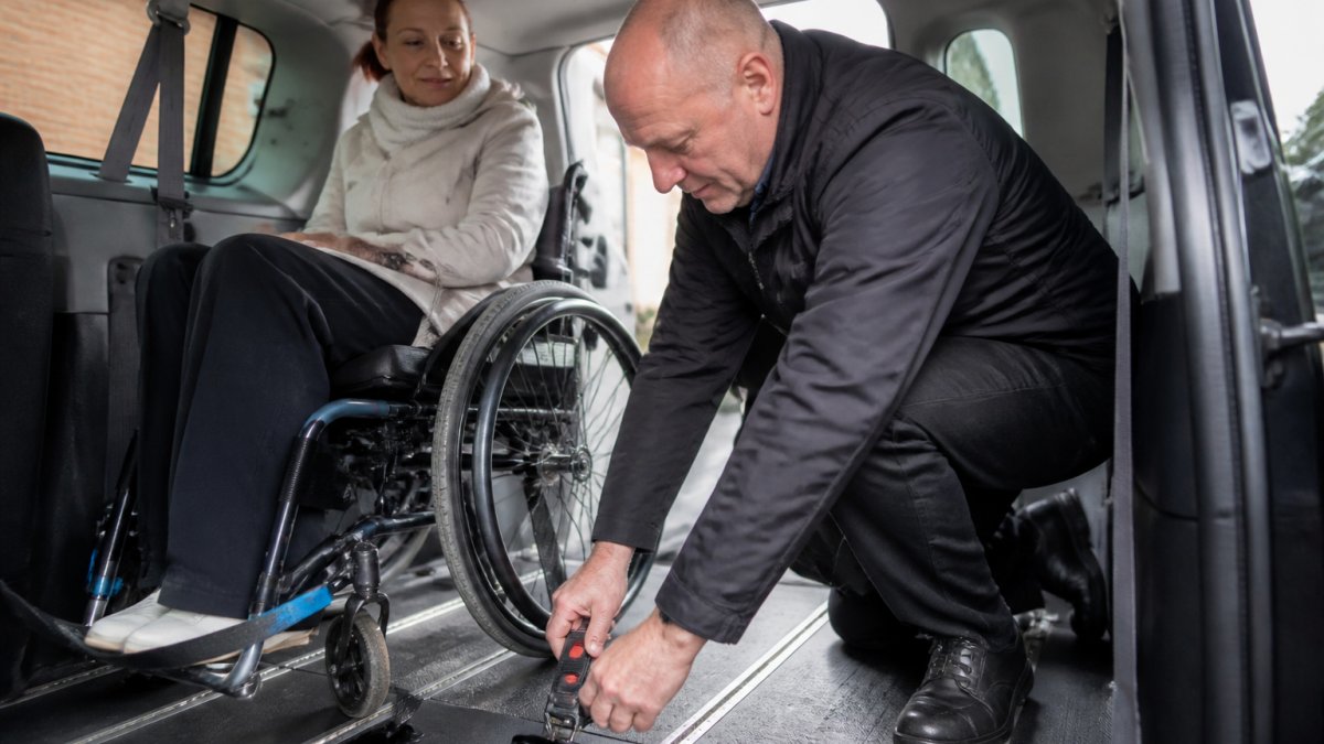Een ervaren chauffeur in een rolstoeltaxi die een rolstoel met een bevestigingssysteem vastzet op de vloer van het voertuig, terwijl de passagier rustig toekijkt