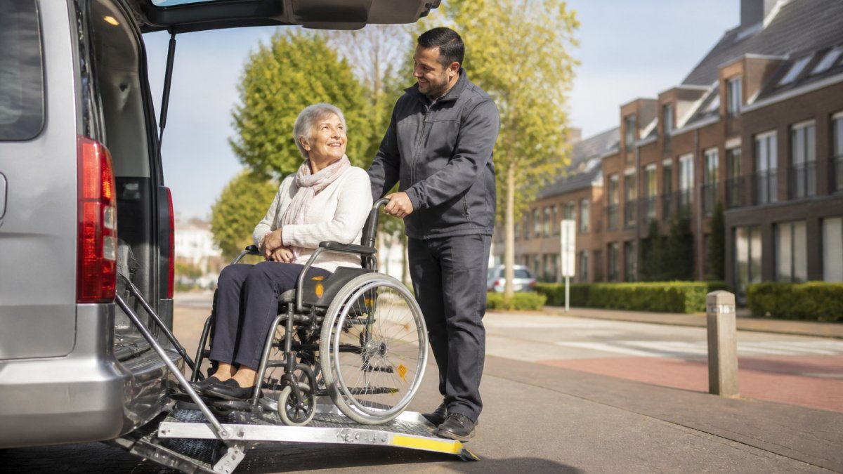 Een oudere mevrouw in een rolstoel wordt door een vriendelijke chauffeur in een rolstoelbus geholpen bij het instappen, met een woonwijk in Utrecht op de achtergrond, zacht ochtendlicht